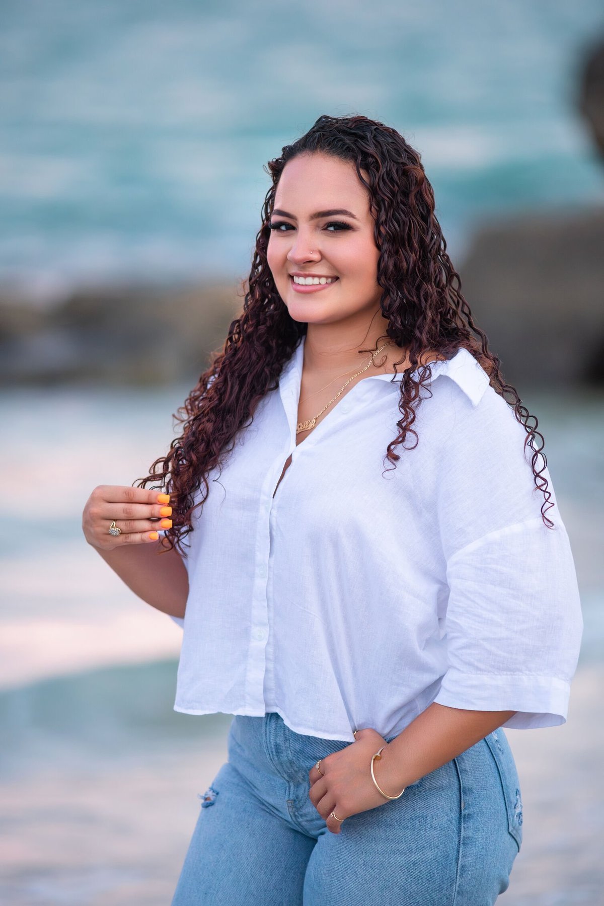 Smiling woman with long curly dark hair wearing a white button-up shirt and jeans, standing on a beach with ocean in background